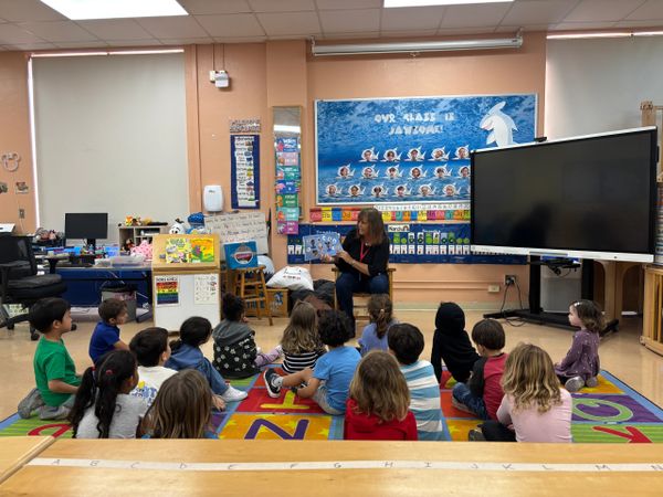 Teacher reading a book to young students seated on a colorful carpet in a classroom.