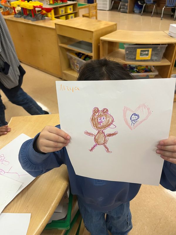 Child proudly shows a colorful drawing of a bear and a heart in a classroom.