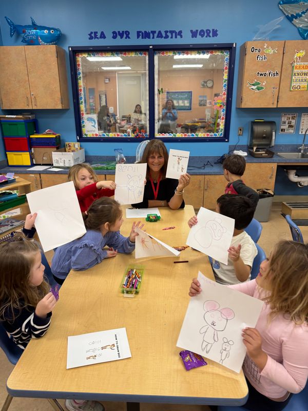 Children proudly show their drawings around a table with their teacher in a colorful classroom.