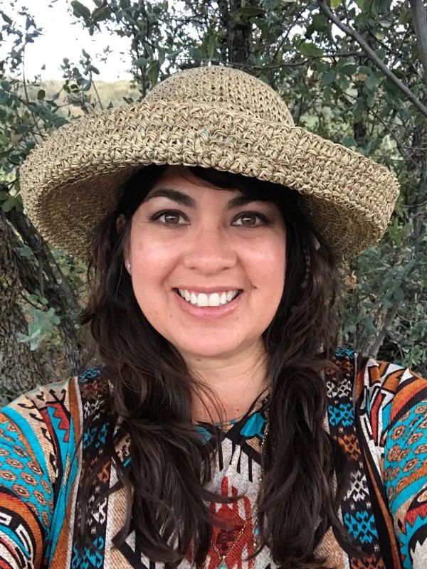 Smiling woman wearing a woven hat and colorful patterned shirt outdoors.