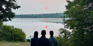 Three people watch a red sun setting over a calm lake surrounded by trees.