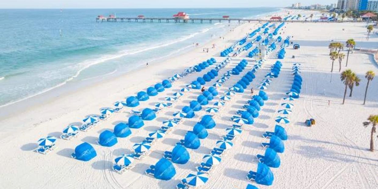 Rows of blue umbrellas and cabanas line a sunny beach near a pier.