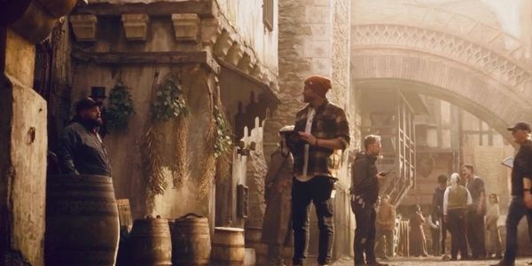 People walking and standing in a rustic alley with barrels and plants.