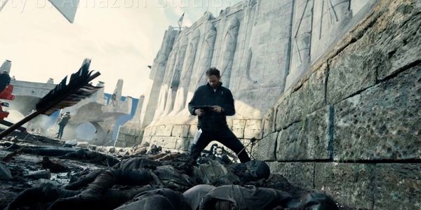 Man stands over fallen soldiers near a large stone wall in a battle scene.