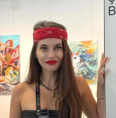 A woman with a red bandana and red lipstick stands smiling in an art gallery.
