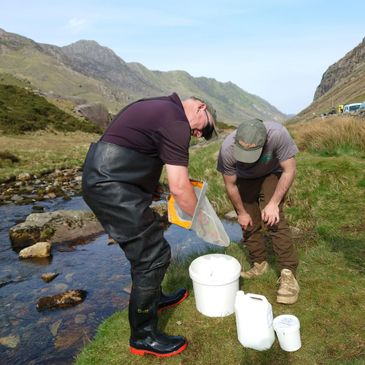 Two men working by a stream with fishing gear in a mountainous area.