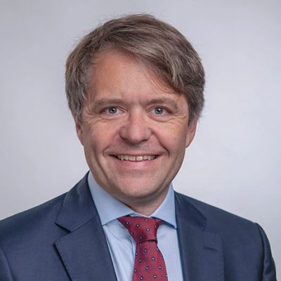 Smiling man in a suit with a red tie against a plain background.