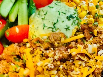 Close-up of a colorful taco salad bowl with ground meat, corn, tomatoes, cucumbers, and green sauce.