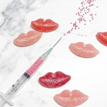 Syringe with pink beads next to sugar-coated lips-shaped gummies on marble.