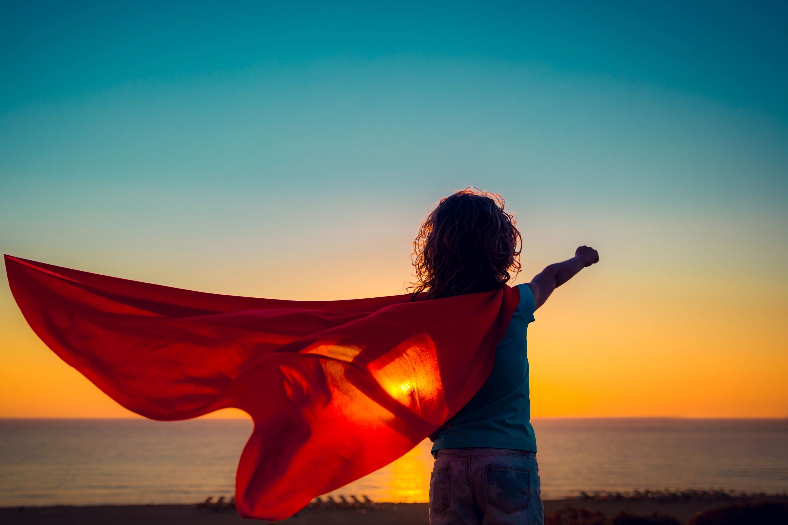 picture of a child wearing a superhero cape overlooking the setting sun over the ocean.