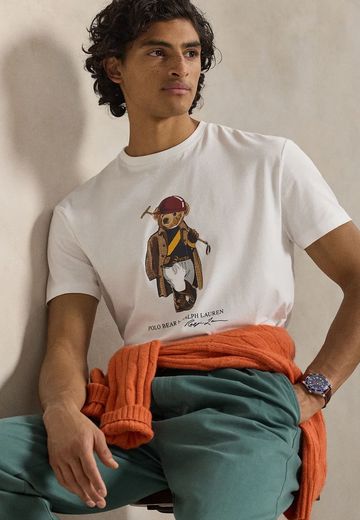Man wearing a white Ralph Lauren Polo Bear t-shirt with green pants and an orange sweater tied around his waist.