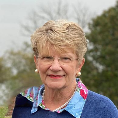 Elderly woman with glasses and pearl jewelry smiling outdoors.