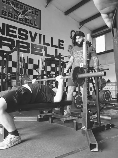Beginner lifter benching while more experienced lifter spots them - friendly, supportive atmosphere.