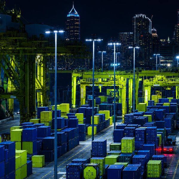 Night view of a brightly lit shipping container yard with city skyline backdrop.