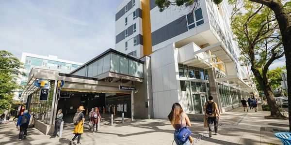 People walking near a modern building and light rail station under a clear sky.