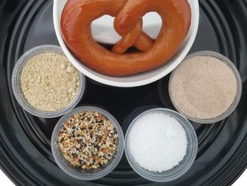 Bakers Mann soft pretzel on plate with four cups of SELECT shaker toppings or dip sauces