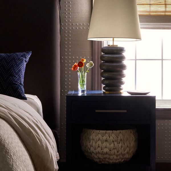 Cozy bedroom corner with a dark nightstand, modern lamp, and small vase of flowers by the window.