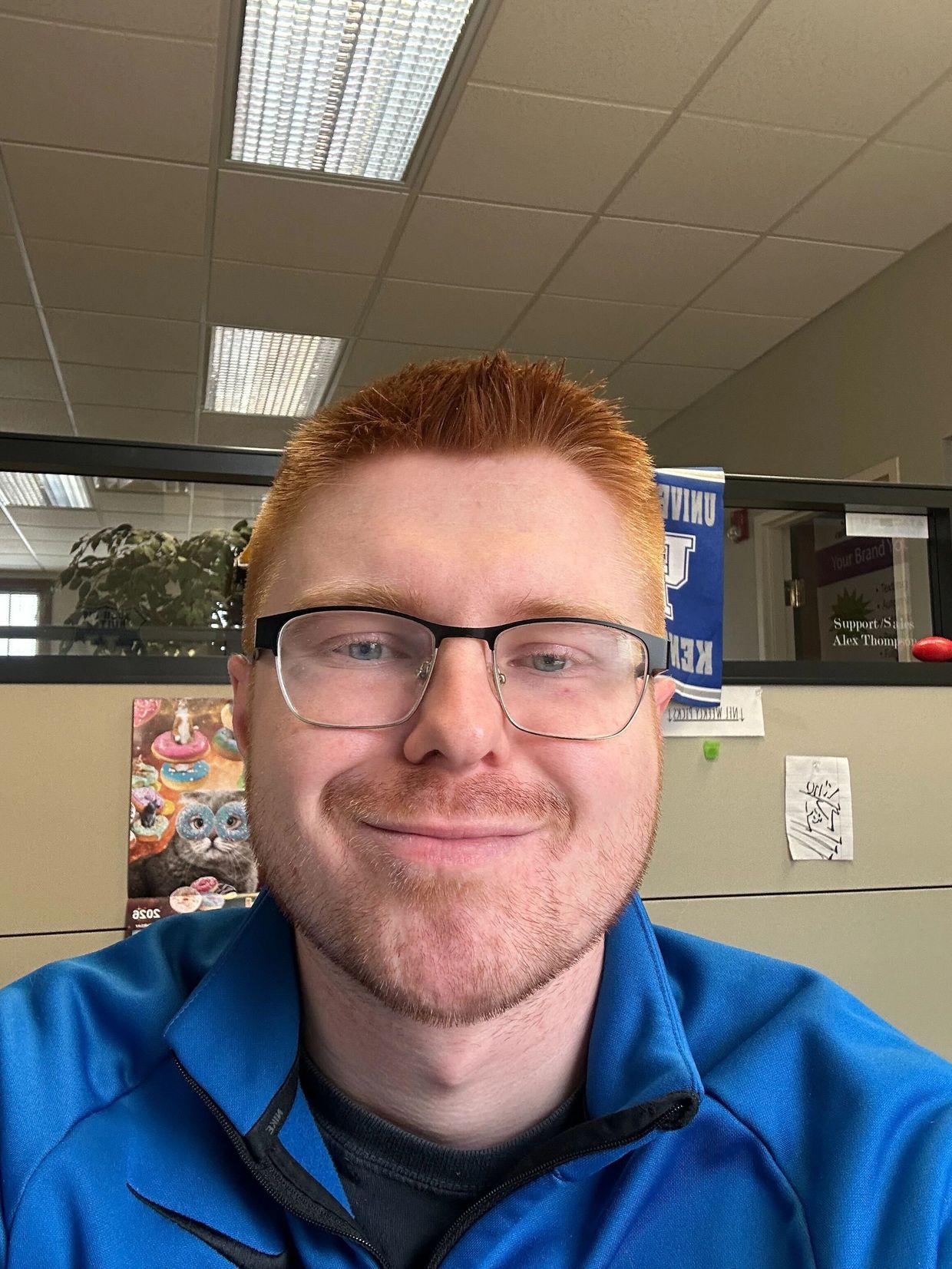 Smiling man with red hair and glasses wearing a blue jacket in an office cubicle.