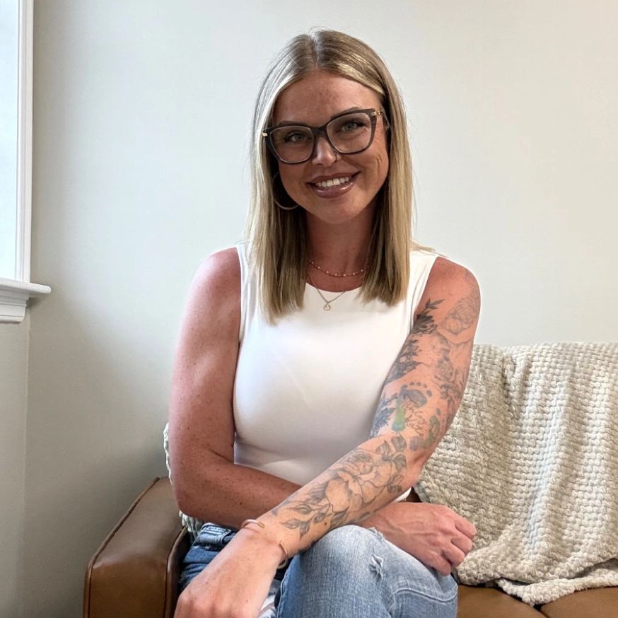 Smiling woman with glasses and sleeve tattoo sitting relaxed.