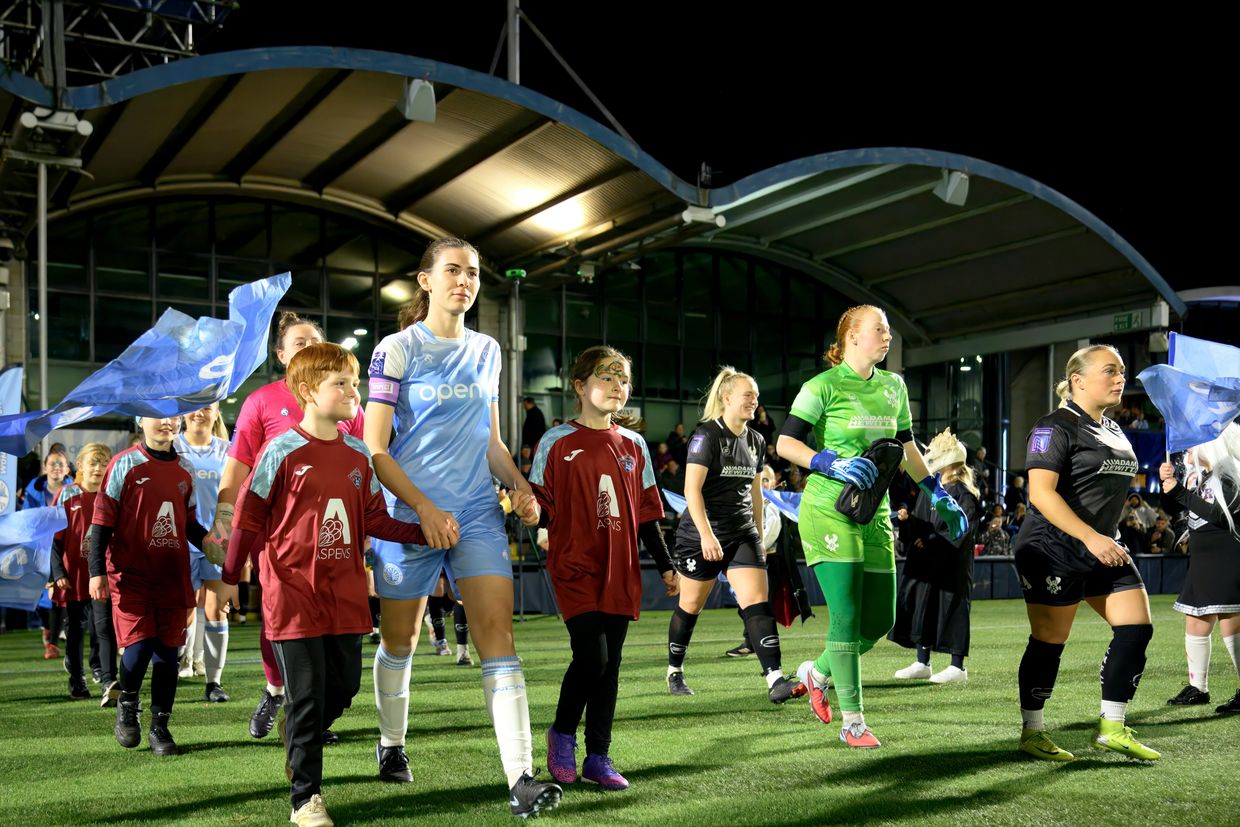 Women's soccer teams enter the field with young mascots at night under stadium lights.