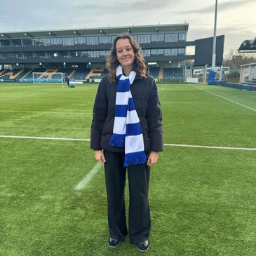 Woman smiling on a soccer field wearing a black jacket and blue-white scarf.