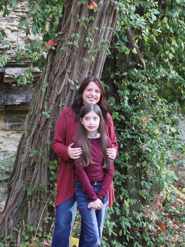 A smiling woman and girl standing in front of a large tree covered with vines.