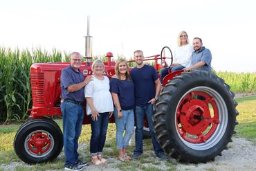 A family poses with a red tractor in a rural setting near a cornfield.
