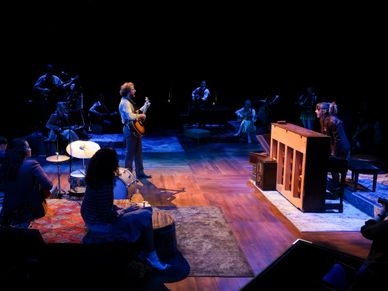 An actor plays guitar facing another actor playing piano.