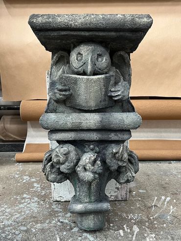 A faux stone sculpture of an owl reading a book, resting on a column capital stands on the ground.