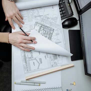 Hands working on architectural blueprints at a desk with computer and drawing tools.