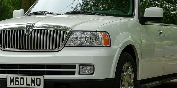 White luxury limousine parked on a paved driveway with green trees in the background.
