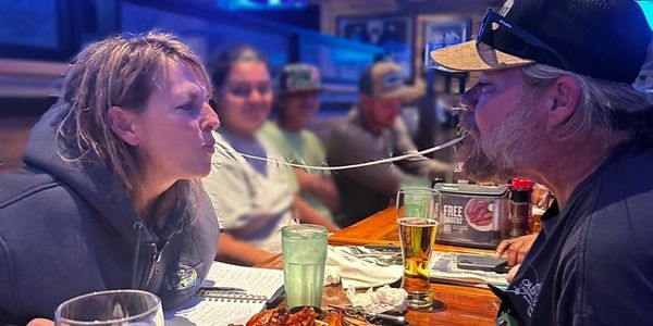 Two people sharing a long noodle at a restaurant with friends in the background.
