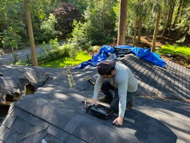 A worker repairs a roof surrounded by trees and greenery.