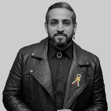 Man in black leather jacket with a ribbon pin, posing confidently against a gray background.