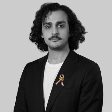 Man with curly hair and mustache wearing a black blazer and white shirt.