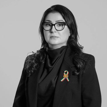 Serious woman wearing glasses and a black blazer with a colorful ribbon pin.