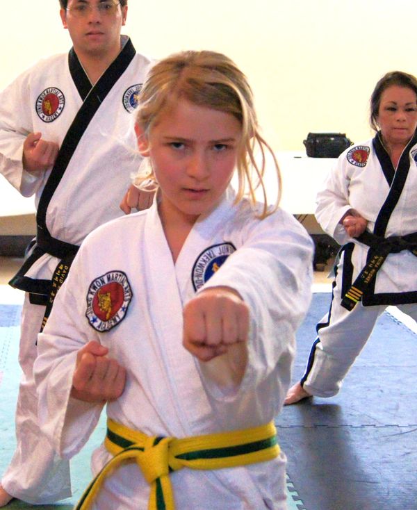 Close-up of a young blonde girl in a white TKD uniform, punching toward the camera.