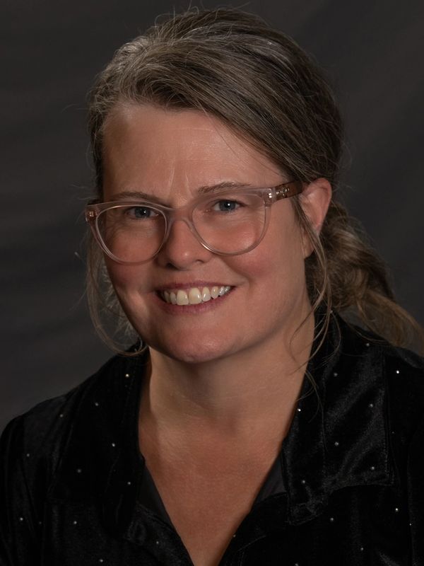 Smiling woman with glasses and tied-back hair in dark clothing.