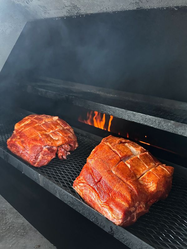 Two large pieces of seasoned meat smoking on a grill with visible flames.
