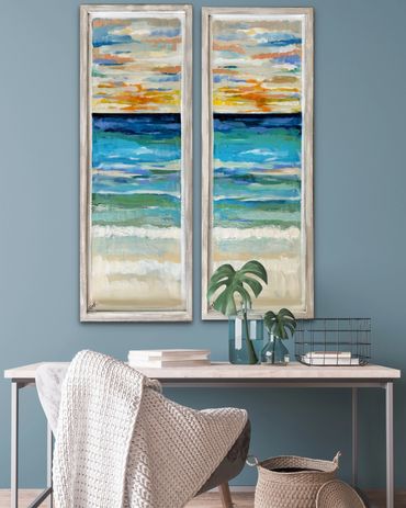 Coastal-themed diptych paintings above a minimalist desk with plants and a cozy chair.