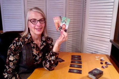 A woman sitting at a desk holding up two tarot cards with other tarot cards on the desk in a spread.