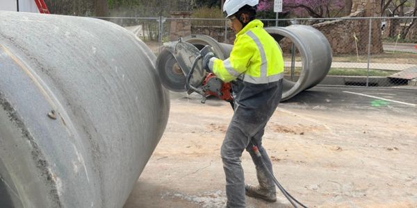 Worker cutting large concrete pipe with power saw at construction site.