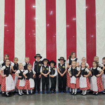 Children in traditional folk costumes pose before a large American flag backdrop.