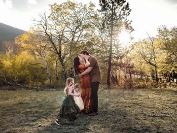 Couple embracing in a sunlit forest clearing with two children playing nearby.