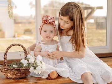 Two young girls in white dresses sitting with baskets and flowers.