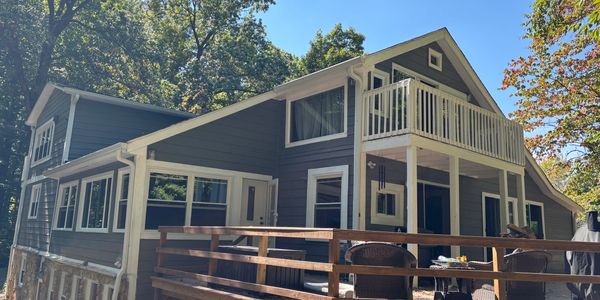Gray house with white trim surrounded by trees under a clear blue sky.