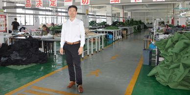 Man standing in a garment factory with sewing workstations and fabric piles.