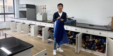 Man holding blue fabric sheet in a lab with open cabinets filled with colorful rolls.