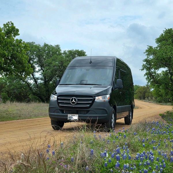 A black Mercedes van drives on a dirt road lined with vibrant bluebonnets