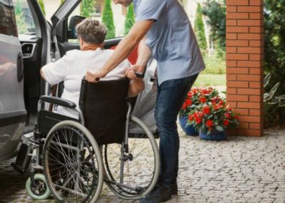 A man helps an elderly person in a wheelchair into a vehicle.